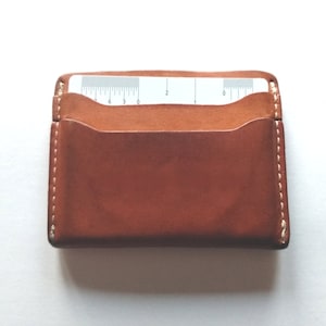 Pu&ograve; includere: Un porta carte in pelle marrone con un bordo cucito bianco. Il porta carte ha una tasca per una carta con un righello stampato su di essa.