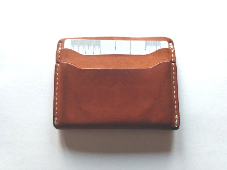 Pu&ograve; includere: Un porta carte in pelle marrone con un bordo cucito bianco. Il porta carte ha una tasca per una carta con un righello stampato su di essa.