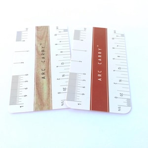 May include: Two rectangular pocket-sized measuring cards with rounded corners. One card features a light wood grain center stripe, the other a solid rust-colored stripe. Both display the text ARC CARRY, imperial and metric markings.