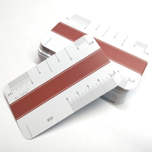 May include: A stack of rectangular white cards with rounded corners, featuring a horizontal terracotta-colored stripe across the center. Each card includes printed ruler markings in inches, numerical labels, and the text Arc Carry and USA.