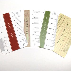 May include: Four rectangular pocket rulers with rounded corners, featuring various designs including a red stripe with ARC CARRY text, a wood grain pattern, a green stripe with CIVIL CARRY 808 text, and a light wood finish labeled Ruled By The Ruler.