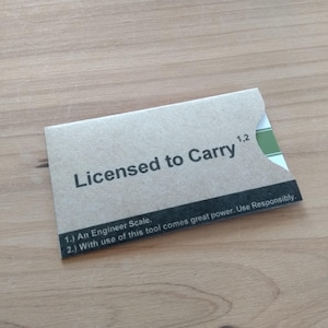 May include: A small, rectangular, kraft paper card with the text "Licensed to Carry" in black. The card also includes the text "1,2" and two numbered statements in black. The card is on a wooden surface.