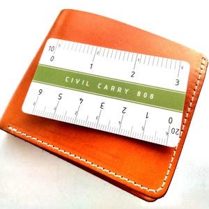 May include: A smooth orange leather bifold wallet with white stitching. A white and green rectangular ruler card is placed on top, featuring the text CIVIL CARRY 808 and markings in inches up to 3 inches and centimeters up to 7 centimeters.