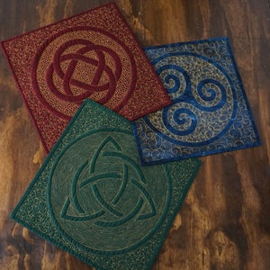 Può includere: Tre quadrati di tessuto decorativi con motivi di nodi celtici. Il quadrato superiore è rosso con un motivo di nodo dorato. Il quadrato inferiore sinistro è verde con un motivo di nodo dorato. Il quadrato inferiore destro è blu con un motivo a spirale dorato.