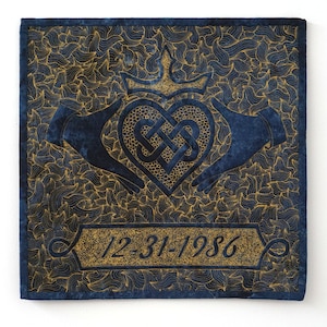 Op de afbeelding: Een blauw gestikt vierkant van stof met een gouden geborduurd ontwerp van een hart met een Keltische knoop erin. Het hart wordt vastgehouden door twee handen. De datum "12-31-1986" is onder het hart geborduurd.