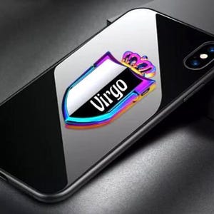 May include: Black smartphone with a reflective surface. A colorful, iridescent shield-shaped phone grip is attached, featuring the word "Virgo" in white. The grip is topped with a gold crown.