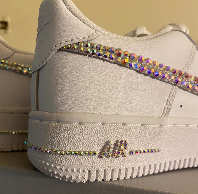 May include: White leather sneakers with iridescent rhinestones on the swoosh and around the "AIR" logo on the sole.