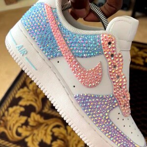 May include: White leather low-top sneakers with a pink, blue, and iridescent crystal design. The sneakers have a white rubber sole with the word "AIR" printed on the side.