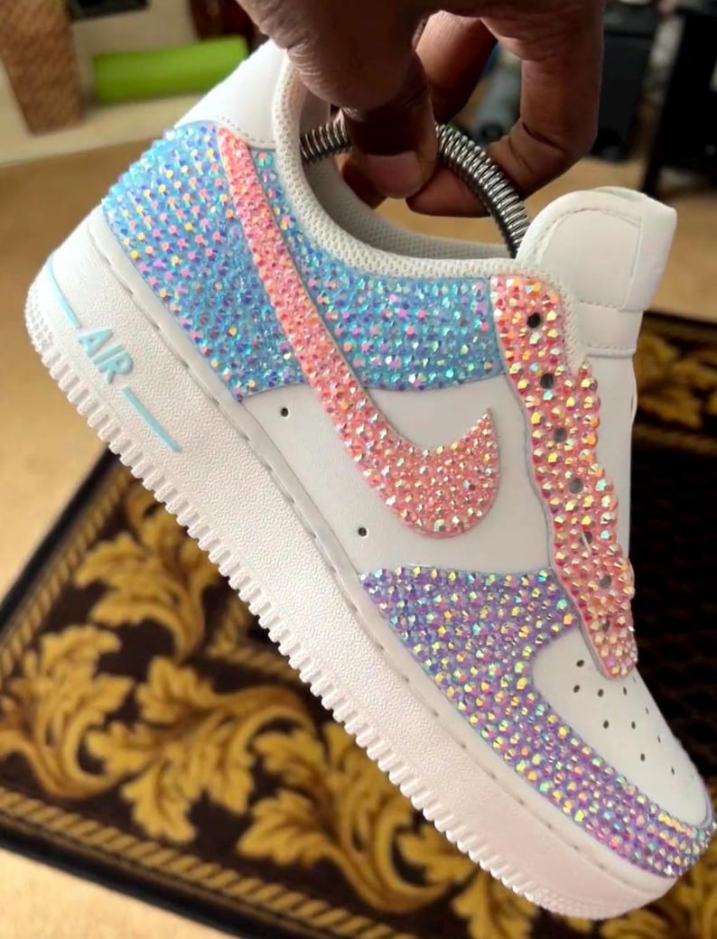 May include: White leather low-top sneakers with a pink, blue, and iridescent crystal design. The sneakers have a white rubber sole with the word "AIR" printed on the side.