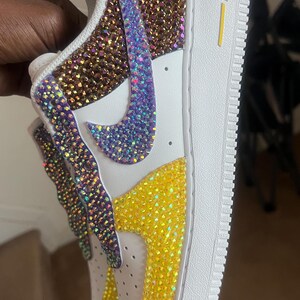 May include: White leather sneakers with a purple, gold, and yellow rhinestone design. The sneakers have a yellow "AIR" logo on the side.