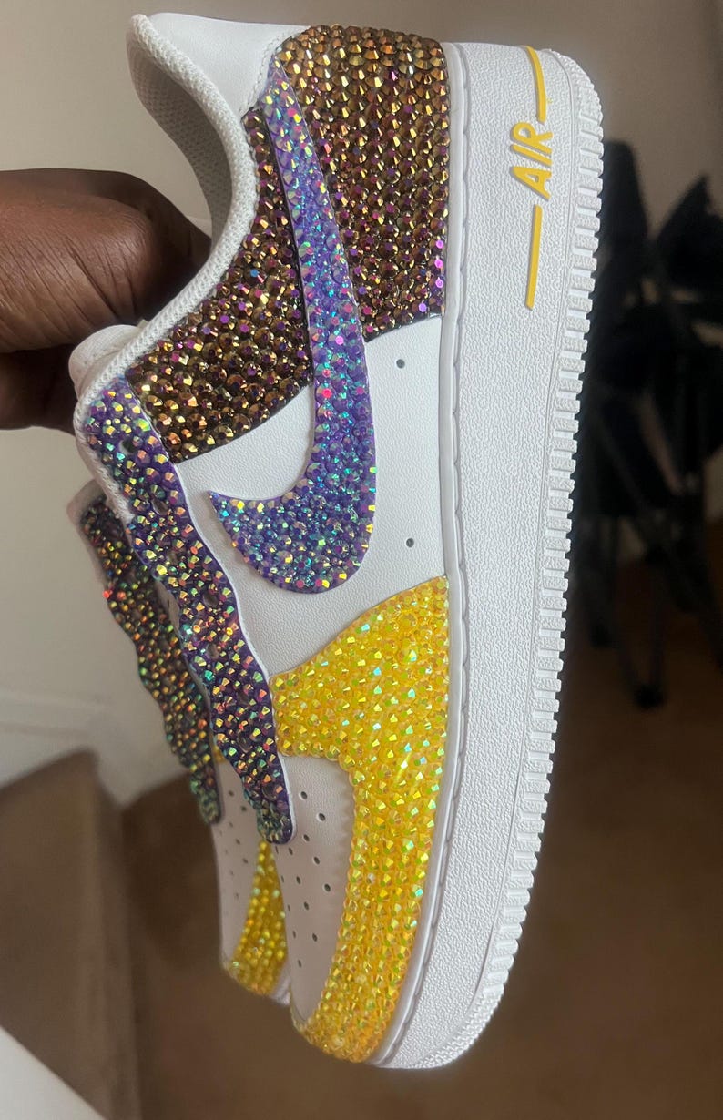 May include: White leather sneakers with a purple, gold, and yellow rhinestone design. The sneakers have a yellow "AIR" logo on the side.