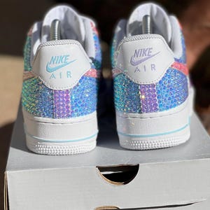 May include: A pair of white Nike Air Force 1 sneakers with a multi-colored iridescent design. The sneakers are covered in rhinestones and have the Nike Air logo on the back.