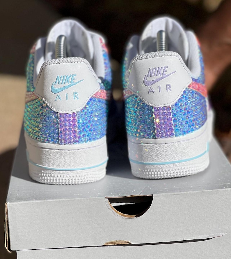 May include: A pair of white Nike Air Force 1 sneakers with a multi-colored iridescent design. The sneakers are covered in rhinestones and have the Nike Air logo on the back.