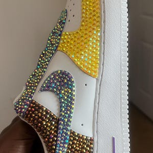 May include: White leather sneakers with a yellow, purple, and brown iridescent rhinestone design. The sneakers have a white sole and a purple "AIR" logo on the back.
