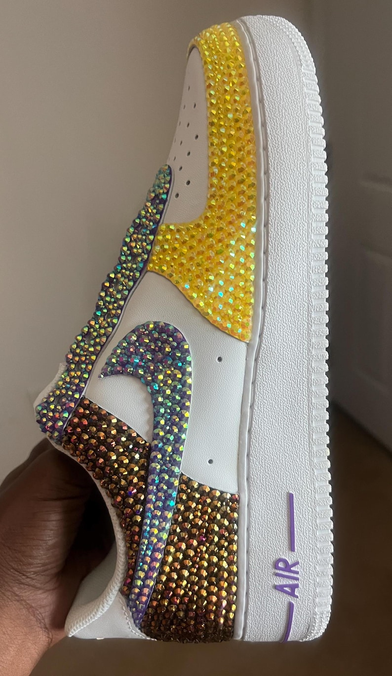 May include: White leather sneakers with a yellow, purple, and brown iridescent rhinestone design. The sneakers have a white sole and a purple "AIR" logo on the back.