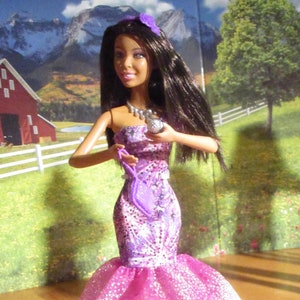 Puede incluir: Una muñeca con un vestido brillante de color rosa y morado con un bolso de mano plateado. La muñeca tiene el pelo largo y negro y lleva una flor morada en el pelo.