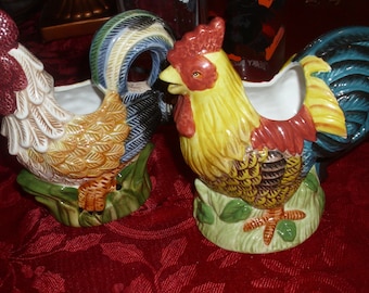 May include: Two ceramic rooster-shaped planters. One rooster is primarily yellow with red and brown accents, the other is cream and brown. Both have a green base with leaves and a blue and white tail. They are decorative and functional.
