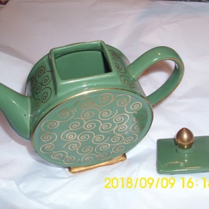 May include: A green teapot with a gold swirl design and a matching lid. The teapot has a square opening on top and a round base.