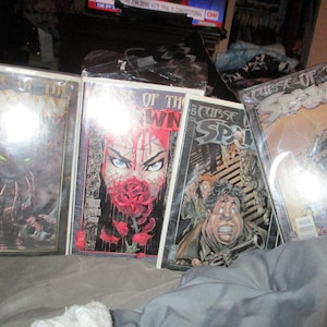 Puede incluir: Cuatro cómics con el título "Curse of the Spawn" en la portada. Las portadas presentan diferentes ilustraciones del personaje Spawn, incluyendo un primer plano de la cara de una mujer con una rosa en la boca, un hombre con una pistola y una criatura con garras.