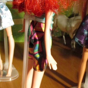 Puede incluir: Una mu&ntilde;eca con cabello rojo brillante y una falda estampada con patrones geom&eacute;tricos coloridos. La mu&ntilde;eca est&aacute; de pie sobre un soporte de pl&aacute;stico transparente. La imagen est&aacute; tomada desde un &aacute;ngulo bajo, con la espalda de la mu&ntilde;eca hacia la c&aacute;mara.
