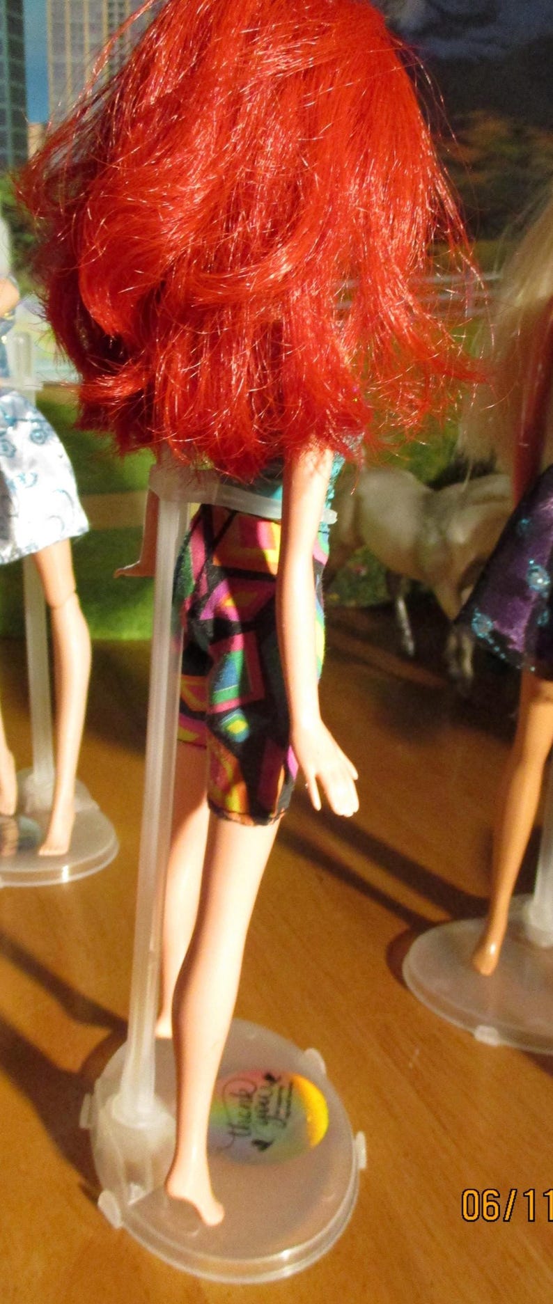 Puede incluir: Una mu&ntilde;eca con cabello rojo brillante y una falda estampada con patrones geom&eacute;tricos coloridos. La mu&ntilde;eca est&aacute; de pie sobre un soporte de pl&aacute;stico transparente. La imagen est&aacute; tomada desde un &aacute;ngulo bajo, con la espalda de la mu&ntilde;eca hacia la c&aacute;mara.