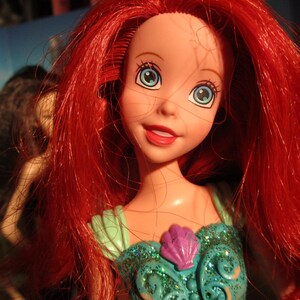 Puede incluir: Mu&ntilde;eca de un personaje con cabello rojo brillante y ojos azules. La mu&ntilde;eca lleva un vestido verde azulado con un detalle de concha p&uacute;rpura. La fecha 06/11/2023 es visible en la parte inferior derecha.