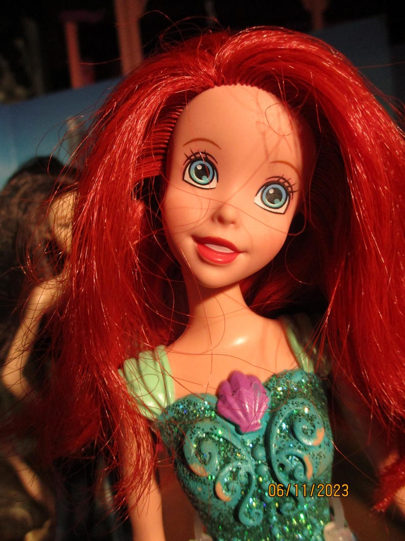 Puede incluir: Mu&ntilde;eca de un personaje con cabello rojo brillante y ojos azules. La mu&ntilde;eca lleva un vestido verde azulado con un detalle de concha p&uacute;rpura. La fecha 06/11/2023 es visible en la parte inferior derecha.