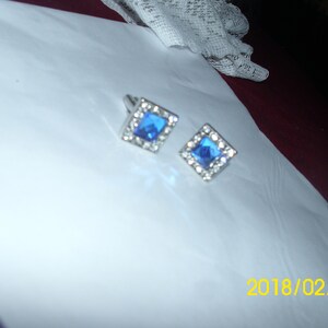 May include: A pair of silver cufflinks with blue square-shaped gemstones surrounded by smaller clear gemstones.