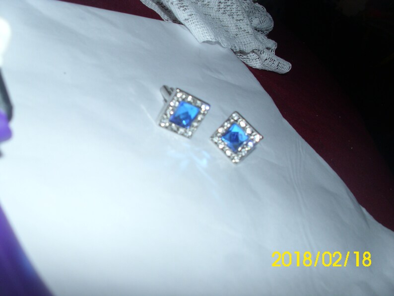May include: A pair of silver cufflinks with blue square-shaped gemstones surrounded by smaller clear gemstones.