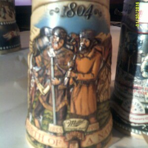 May include: A ceramic beer stein with a brown, tan, and blue design. The stein features a scene of two men in medieval garb, with the text "1804" and "Birth of a Nation" on the front. The Miller Brewing Company logo is also visible.