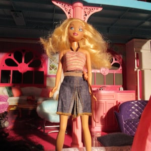 May include: A doll with blonde hair, wearing a striped halter top and a denim skirt. The doll is standing in front of a pink and blue dollhouse. The date 15/04/2023 is visible in the bottom right corner.