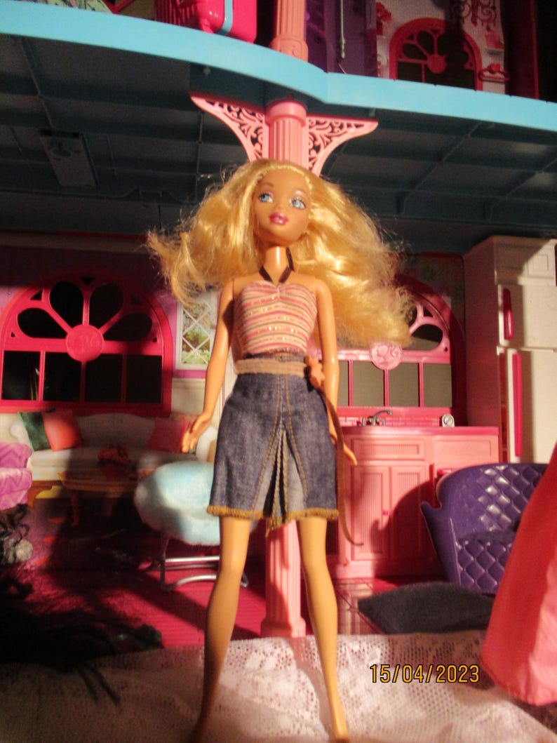 May include: A doll with blonde hair, wearing a striped halter top and a denim skirt. The doll is standing in front of a pink and blue dollhouse. The date 15/04/2023 is visible in the bottom right corner.