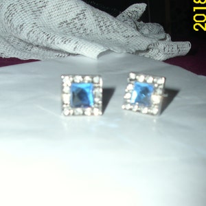 May include: A pair of silver cufflinks with square blue gemstones surrounded by clear rhinestones.