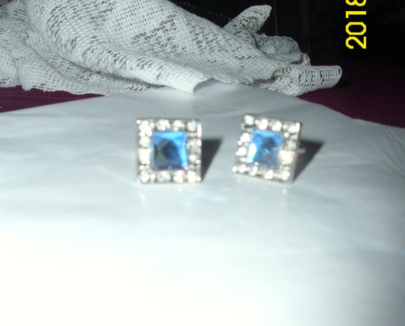 May include: A pair of silver cufflinks with square blue gemstones surrounded by clear rhinestones.