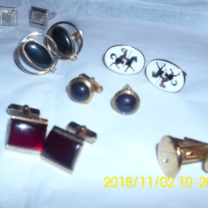 Puede incluir: Una colección de gemelos con varios diseños, incluyendo piedras negras y moradas, piedras rojas y un par con un diseño de caballo y jinete. Los gemelos están sobre una superficie blanca.
