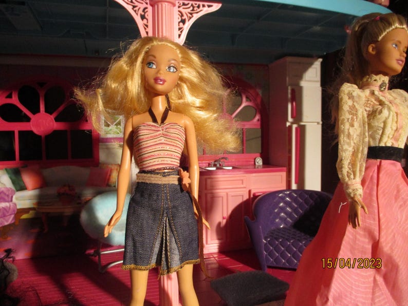 May include: A blonde doll wearing a striped halter top and denim skirt stands in a pink dollhouse. Another doll in a pink dress is visible. The dollhouse has pink furniture and decor.