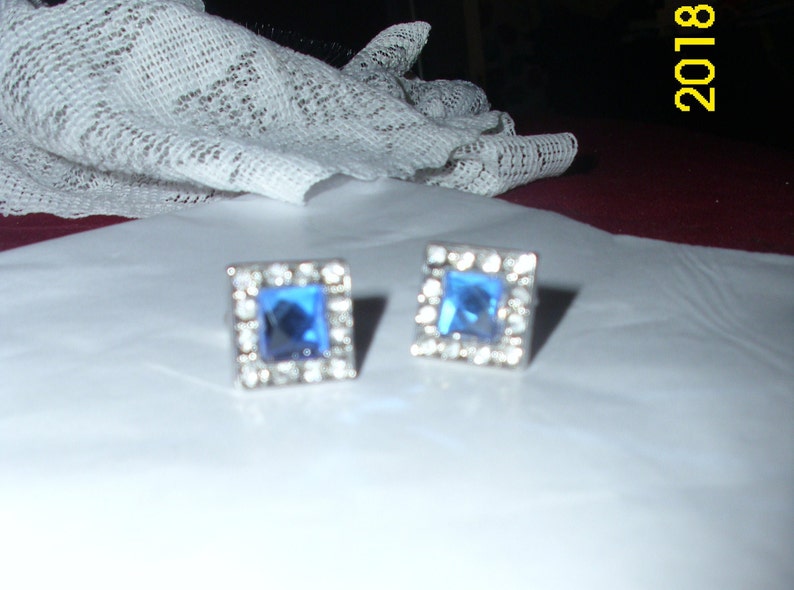 May include: A pair of silver earrings with square blue gemstones surrounded by smaller clear gemstones.