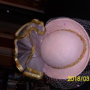 May include: A pink hat with a gold trim and a veil. The hat has a ruffled trim made of a sheer fabric with gold accents.
