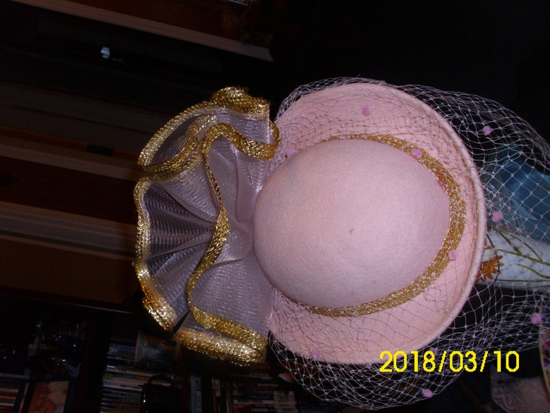 May include: A pink hat with a gold trim and a veil. The hat has a ruffled trim made of a sheer fabric with gold accents.