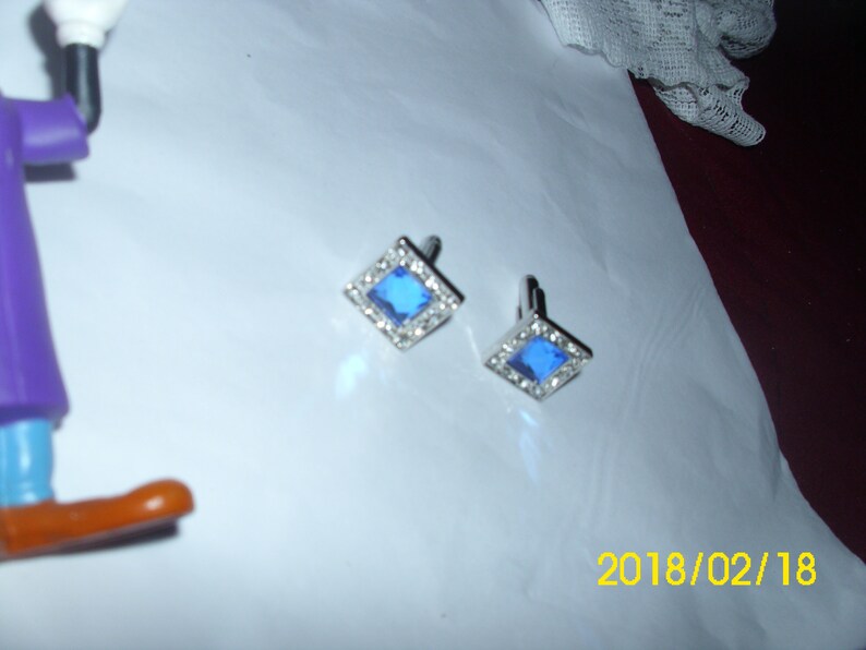 May include: A pair of silver cufflinks with blue square gemstones surrounded by clear rhinestones.