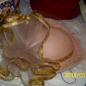 May include: A pink hat with a gold trim and a white tulle bow. The hat is decorated with a gold ribbon and a white tulle bow.