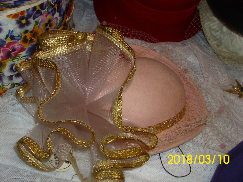 May include: A pink hat with a gold trim and a white tulle bow. The hat is decorated with a gold ribbon and a white tulle bow.