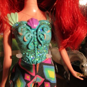 Puede incluir: Mu&ntilde;eca con cabello rojo brillante y un corpi&ntilde;o turquesa adornado con purpurina y una concha morada. El atuendo de la mu&ntilde;eca incluye pantalones estampados coloridos. La fecha 06/11/2023 es visible.
