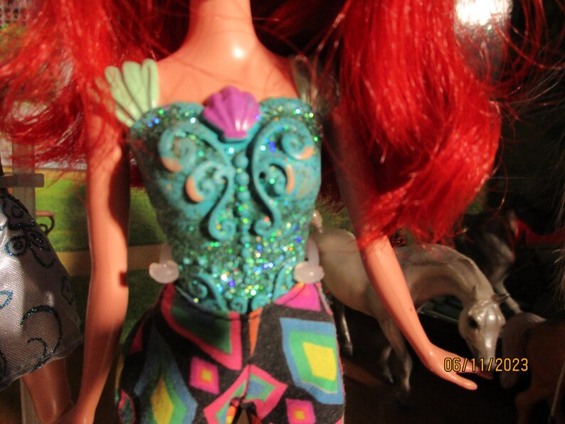 Puede incluir: Mu&ntilde;eca con cabello rojo brillante y un corpi&ntilde;o turquesa adornado con purpurina y una concha morada. El atuendo de la mu&ntilde;eca incluye pantalones estampados coloridos. La fecha 06/11/2023 es visible.