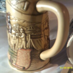 May include: A beige ceramic beer stein with a detailed relief design of a historical scene. The stein has a large handle and a scroll-like design at the bottom. The stein is decorated with brown, green, and white paint.