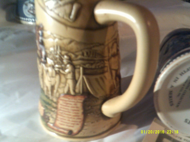May include: A beige ceramic beer stein with a detailed relief design of a historical scene. The stein has a large handle and a scroll-like design at the bottom. The stein is decorated with brown, green, and white paint.