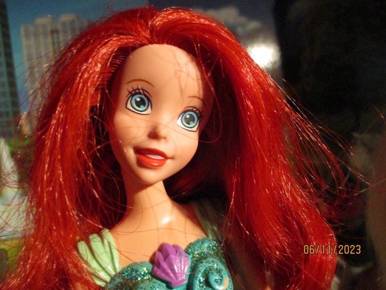 Puede incluir: Mu&ntilde;eca con cabello rojo y ojos azules. La mu&ntilde;eca lleva un atuendo turquesa y morado. La mu&ntilde;eca tiene una expresi&oacute;n sonriente. La fecha 06/11/2023 es visible en la esquina inferior derecha.