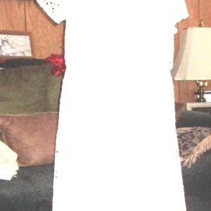 May include: A white short-sleeved dress featuring an intricate eyelet lace pattern throughout the fabric. The garment has a round neckline and a handkerchief hemline that falls to a point, showcasing a delicate, perforated design on the edges.