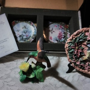 May include: A set of three hummingbird themed porcelain ornaments. The set includes two small round plates with a hummingbird and flowers painted on them, and a larger 3D ornament with a hummingbird perched on a flower. The larger ornament is on a black stand. The set comes with a certificate of authenticity from Bradford Editions.