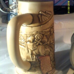 May include: A beige ceramic beer stein with a handle. The stein features a detailed relief design of a scene with people and horses. The design is in shades of brown and beige.